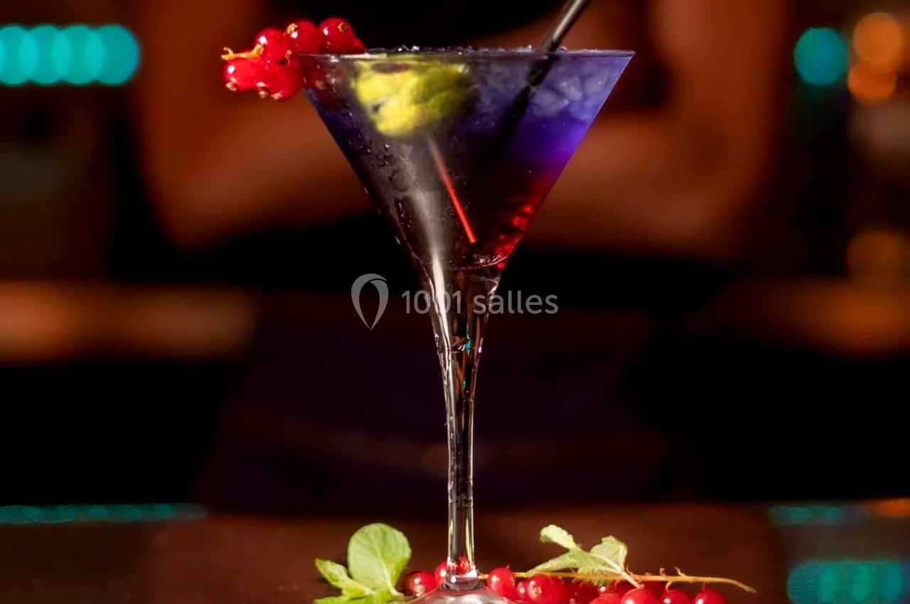 Verre à cocktail violet garni de groseilles rouges, citron vert et menthe, posé sur un comptoir sombre.