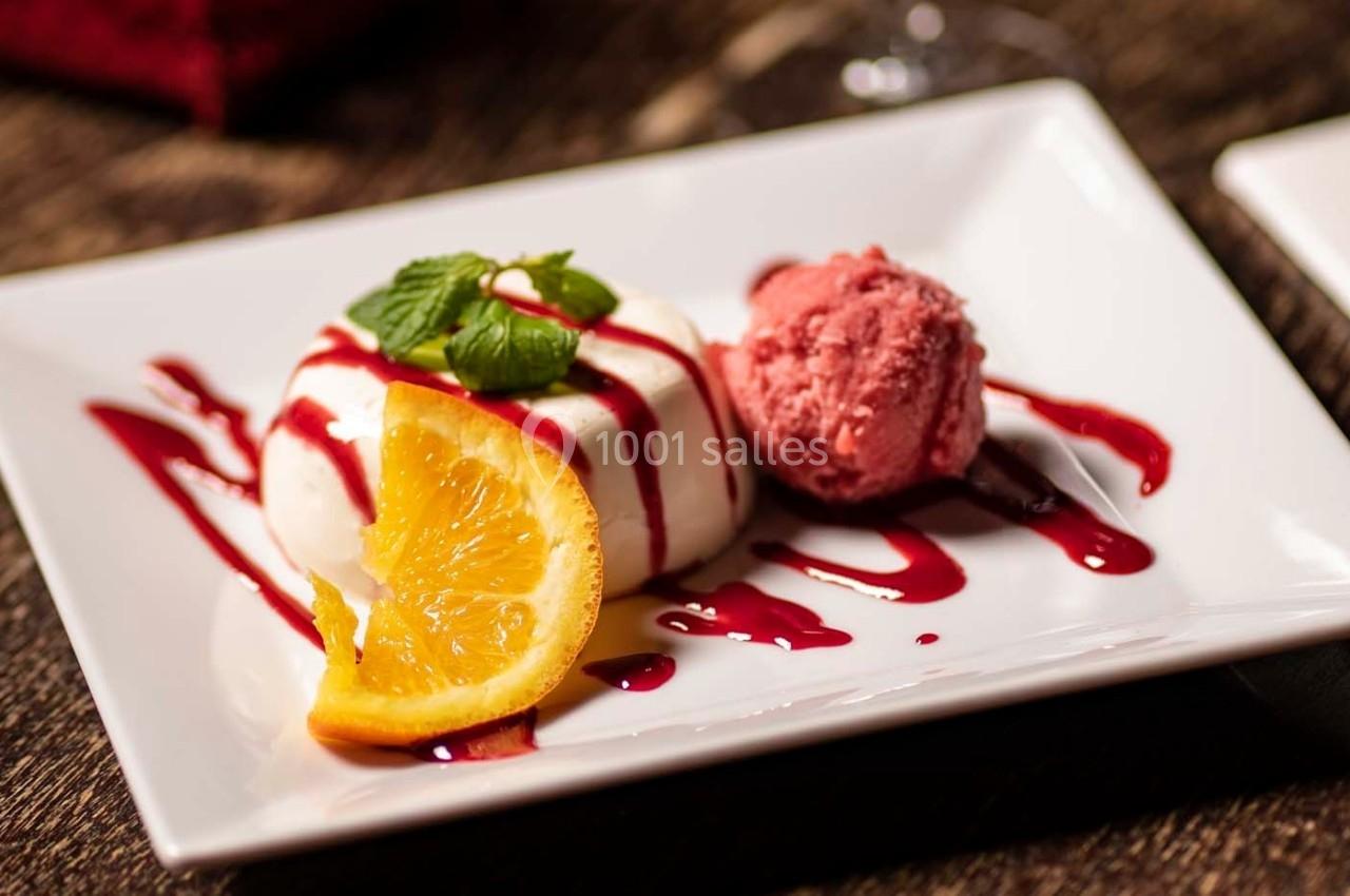 Dessert composé d'une panna cotta garnie de coulis rouge, d'une tranche d'orange, de menthe et d'une boule de glace rose.