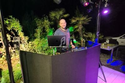 Un DJ mixant en extérieur de nuit, entouré de lumières colorées et de végétation.