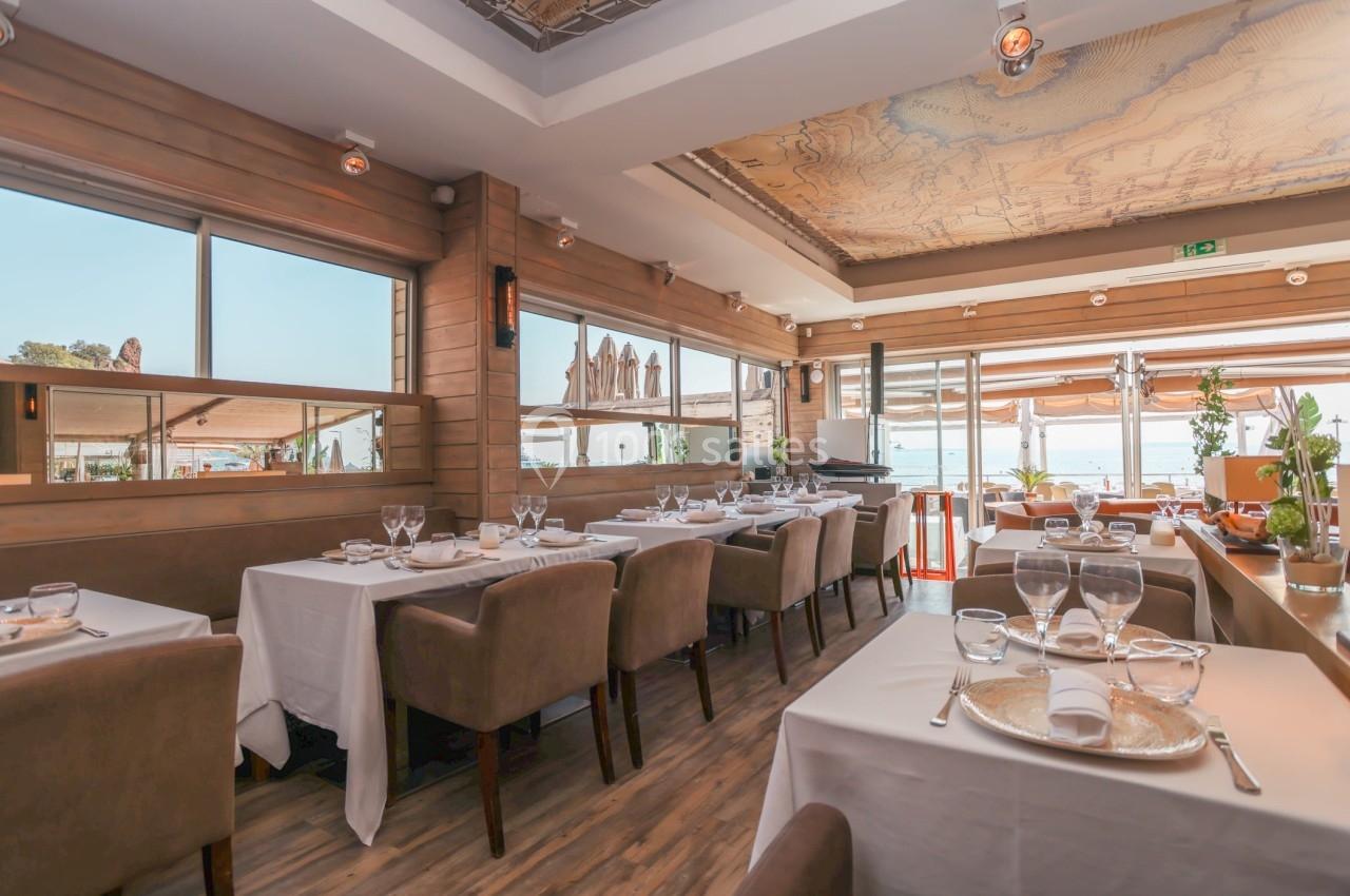 Salle de restaurant lumineuse avec tables dressées, grandes baies vitrées et vue sur l'extérieur.