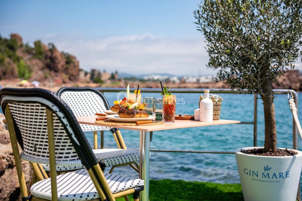 Table en terrasse avec vue sur la mer, plats et cocktails servis, entourée de chaises et d'un olivier en pot.