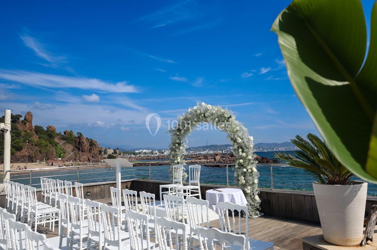 Chaises blanches alignées face à une arche fleurie sur une terrasse en bois avec vue sur la mer et le ciel bleu.