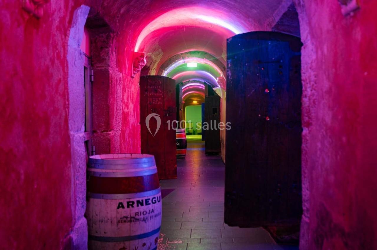 Couloir voûté en pierre éclairé par des lumières colorées, avec des portes en bois ouvertes et un tonneau au premier plan.