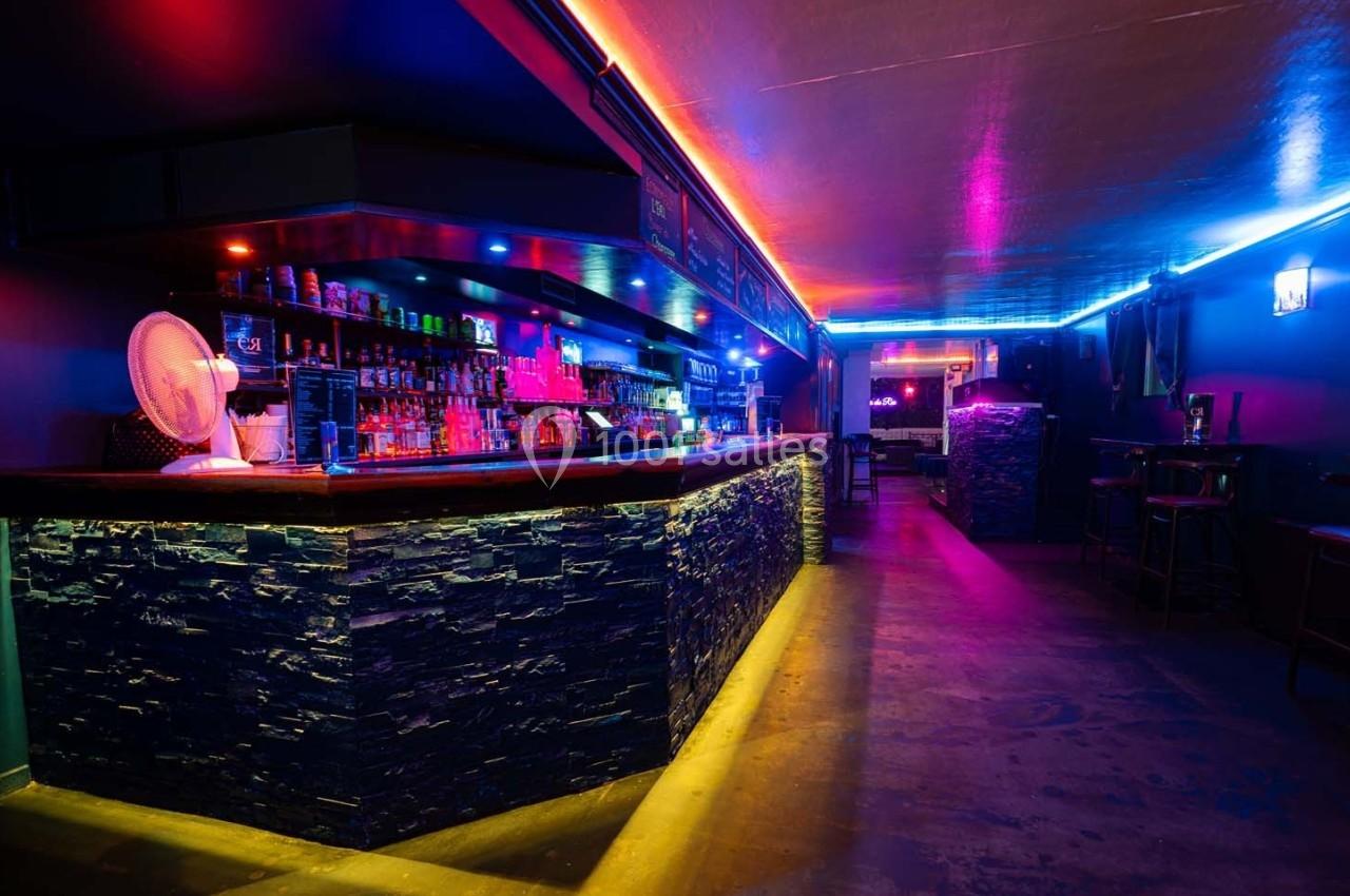 Bar avec comptoir en pierre éclairé par des lumières colorées, étagères remplies de bouteilles et ambiance tamisée.