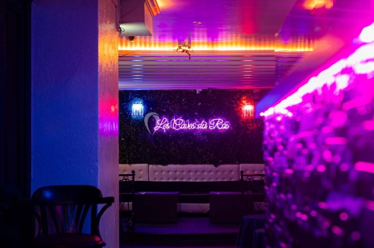 Espace lounge avec banquettes blanches, éclairage néon coloré et enseigne lumineuse ’Les Caves du Roi’.