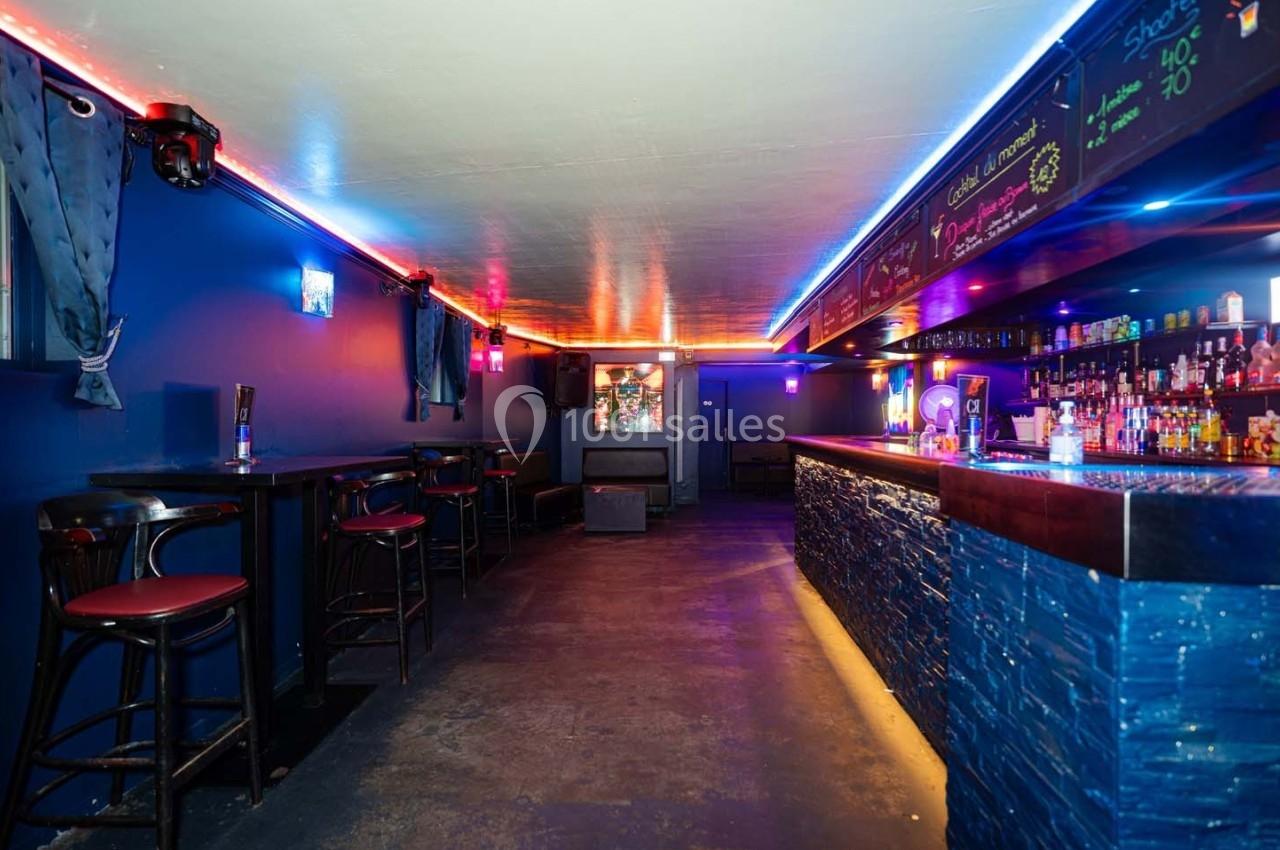 Bar à l'ambiance tamisée avec comptoir éclairé, tabourets hauts et étagères remplies de bouteilles colorées.