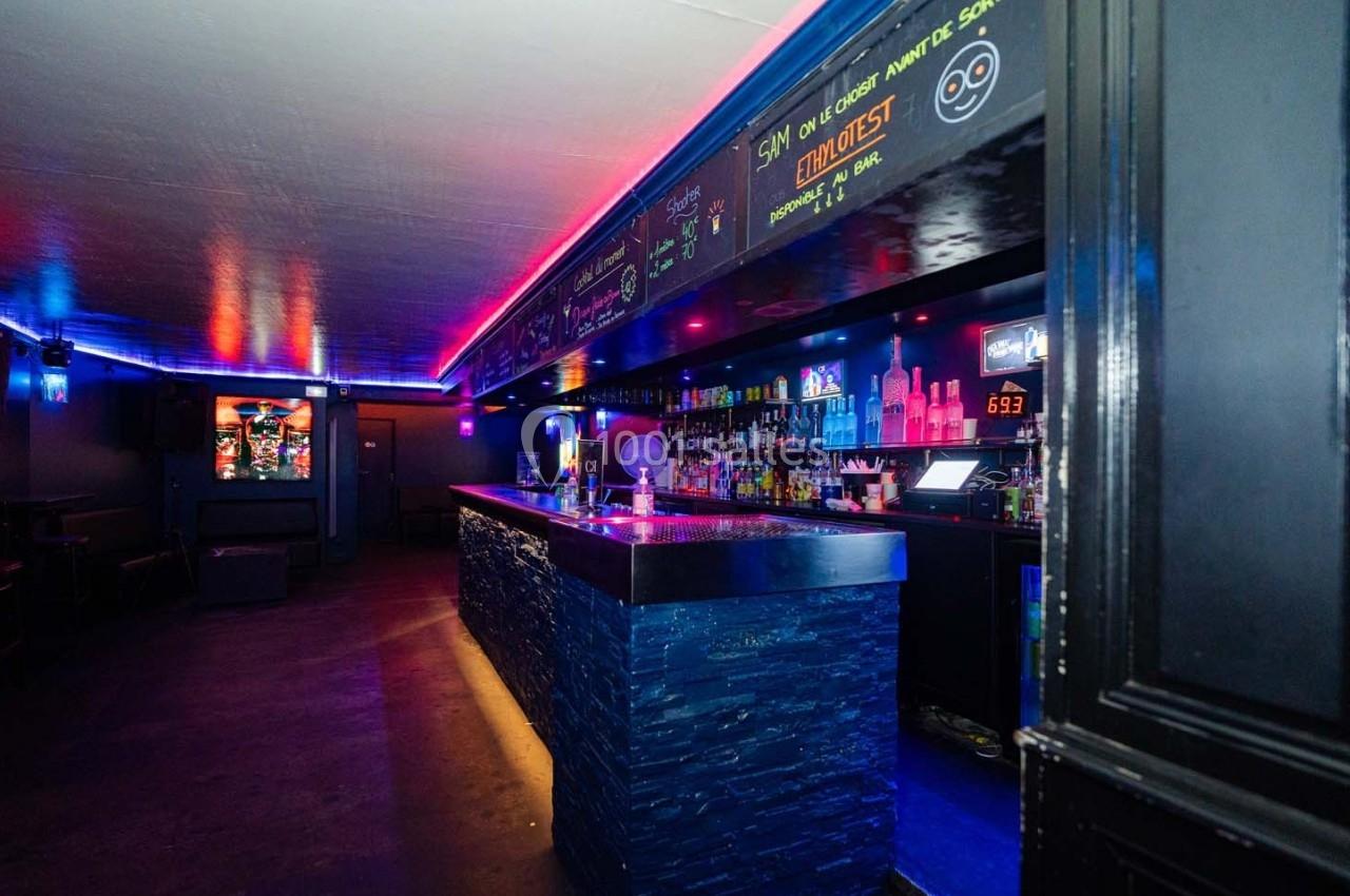 Bar intérieur avec éclairage néon coloré, comptoir en pierre sombre et étagères remplies de bouteilles.