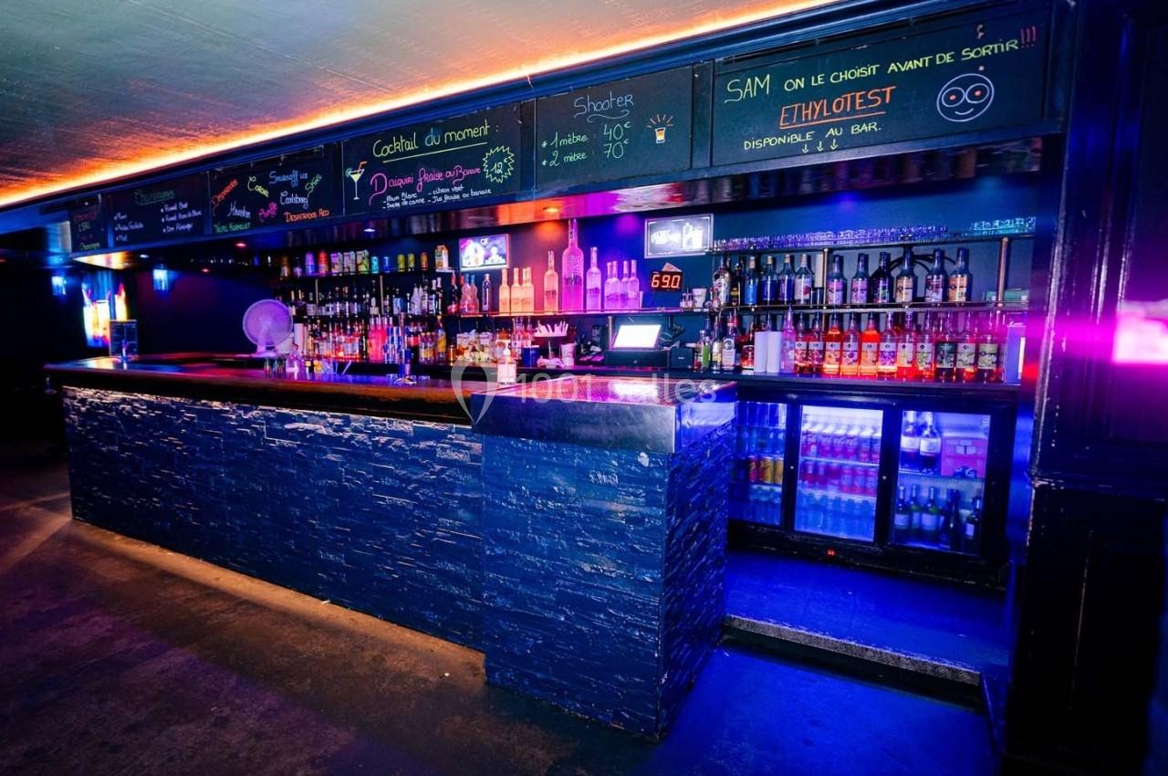 Bar avec comptoir en pierre, étagères remplies de bouteilles, éclairage néon coloré et ardoises avec menus écrits à la craie.