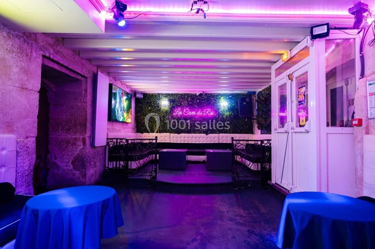 Intérieur d'un bar avec éclairage néon violet, tables bleues, banquettes blanches et murs en pierre.