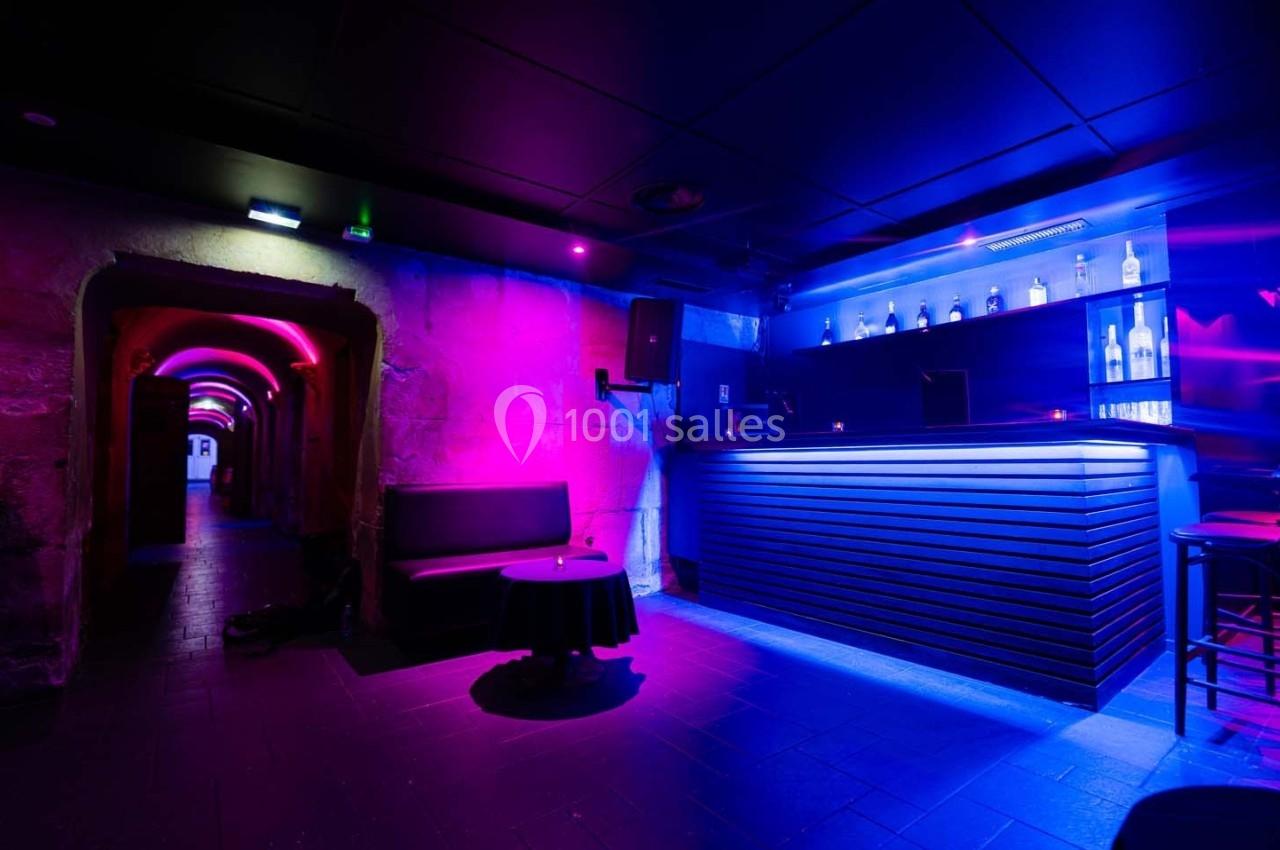 Bar moderne avec éclairage néon violet et bleu, comptoir illuminé, alcôve et sièges dans une ambiance sombre.