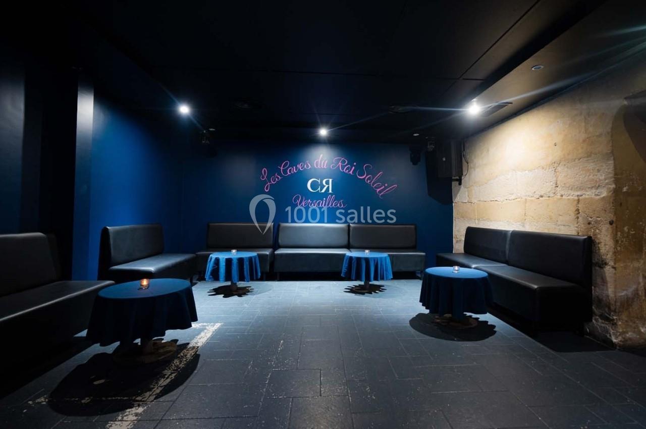Salle sombre avec murs bleus, banquettes noires, petites tables rondes bleues et inscription murale lumineuse.