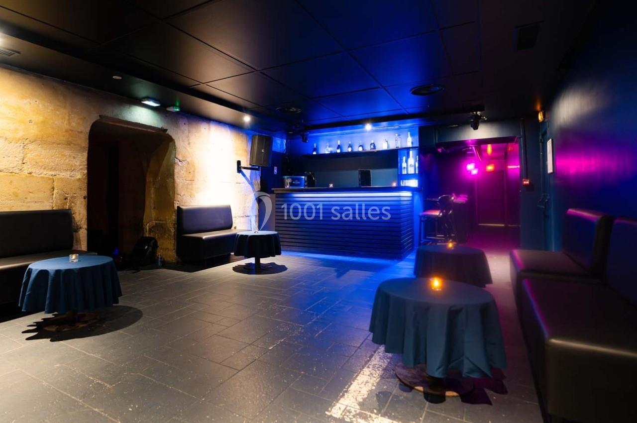 Salle de bar avec éclairage tamisé, tables rondes couvertes de nappes bleues et un comptoir illuminé en arrière-plan.