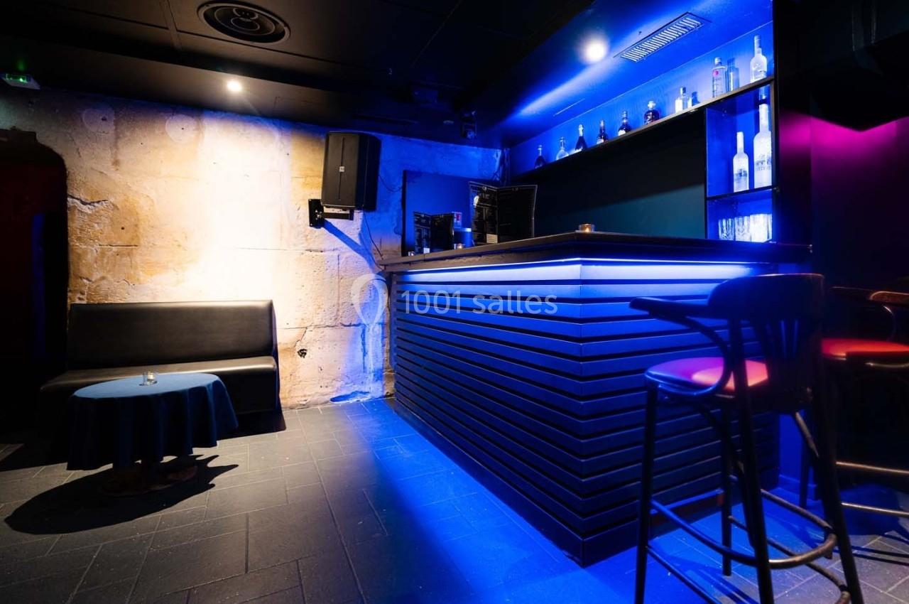 Bar moderne avec éclairage bleu, comptoir en bois sombre, tabourets hauts et banquette contre un mur en pierre.