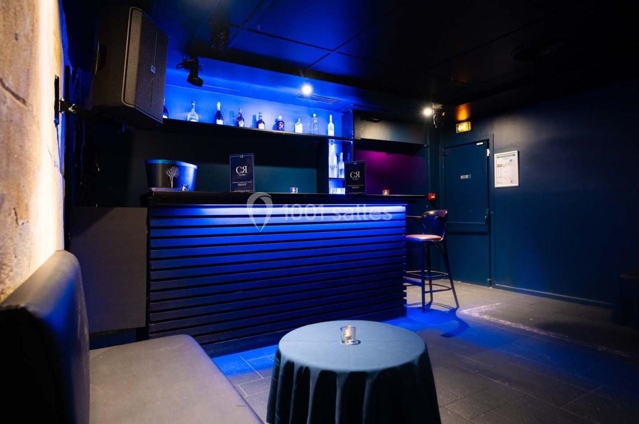 Bar intérieur avec éclairage bleu, comptoir en bois sombre, étagères avec bouteilles et table basse avec bougie.