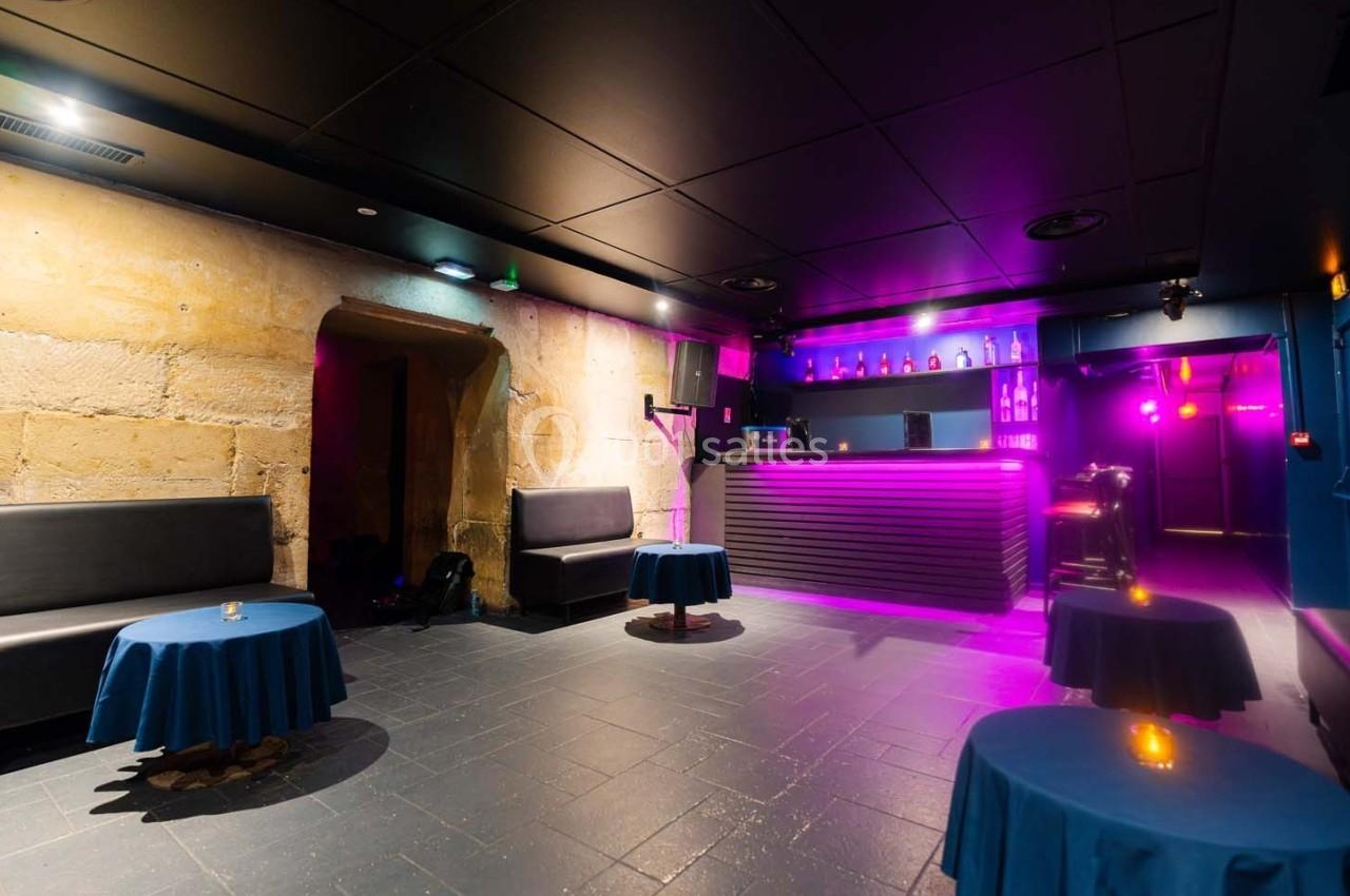 Salle avec murs en pierre, tables basses couvertes de nappes bleues, banquettes et un bar éclairé par des lumières violettes.