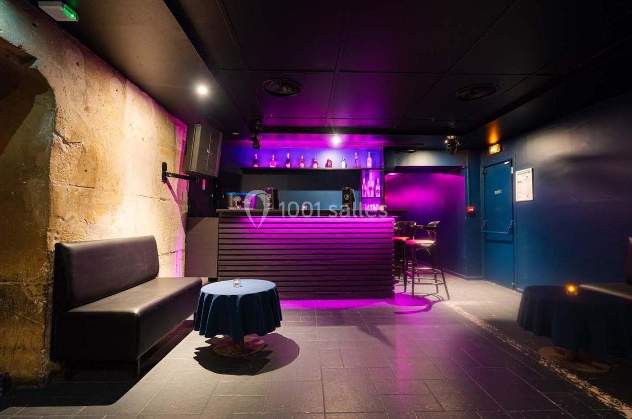 Bar intérieur avec éclairage violet, comptoir en bois, tables basses rondes et banquettes noires sur sol carrelé.