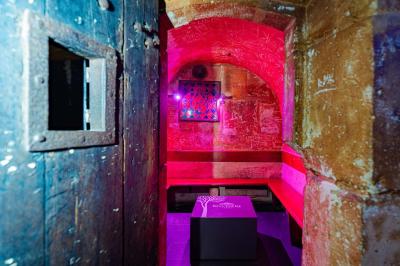 Vue intérieure d'une cellule ancienne avec murs en pierre, bancs en bois et éclairage rose.