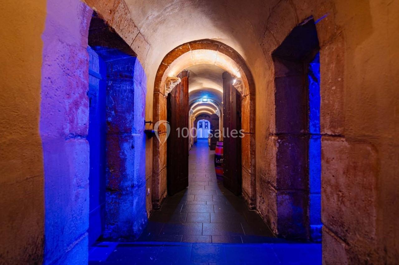 Couloir voûté en pierre avec éclairage bleu, portes en bois ouvertes et sol en dalles sombres.