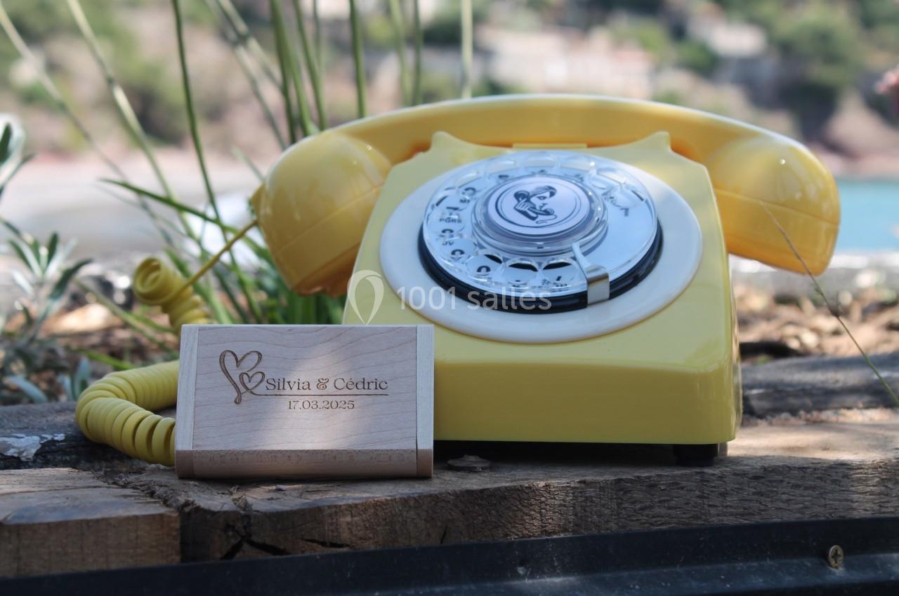 Téléphone jaune rétro posé sur une table en bois à l'extérieur, avec une plaque en bois gravée à côté.