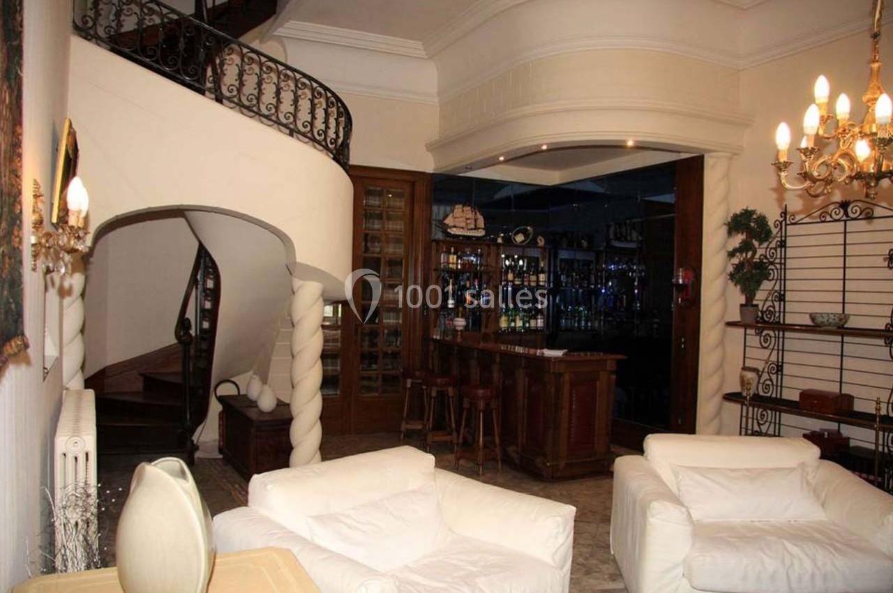 Salon avec fauteuils blancs, escalier en colimaçon, bar en bois et éclairage chaleureux dans un intérieur classique.