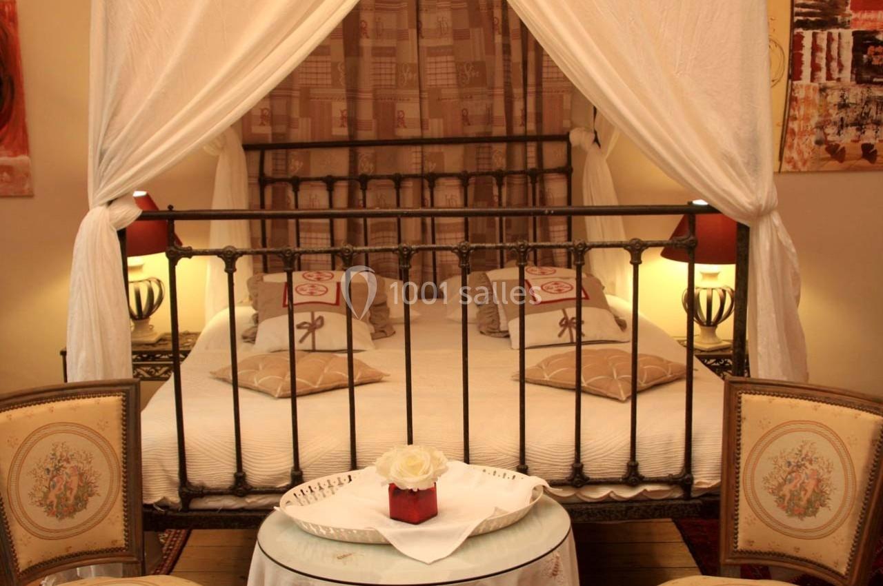 Lit à baldaquin avec draperies blanches, entouré de lampes et fauteuils, dans une chambre décorée chaleureusement.