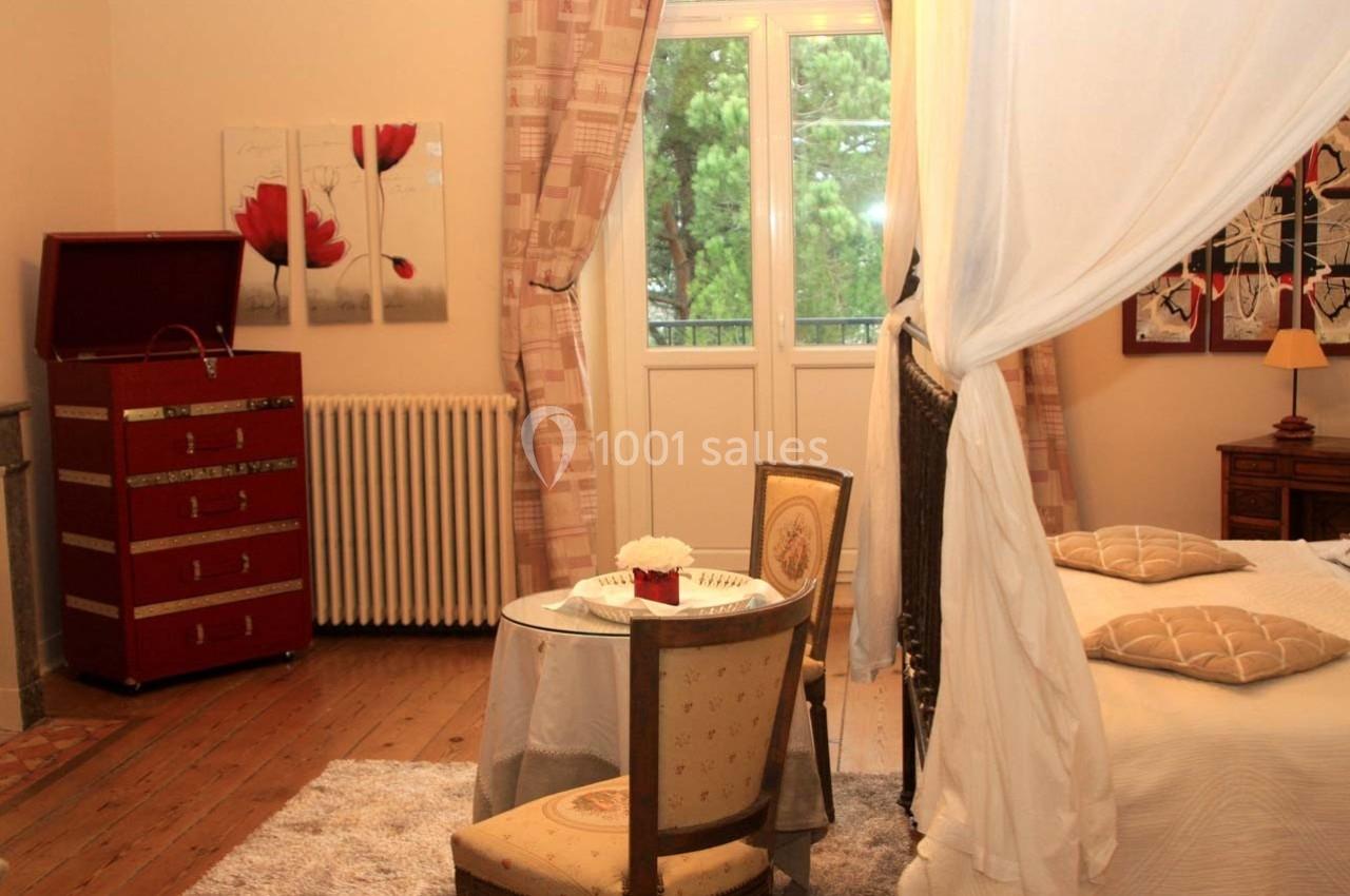 Chambre lumineuse avec lit à baldaquin, table ronde, chaises anciennes, commode rouge et vue sur des arbres par la fenêtre.