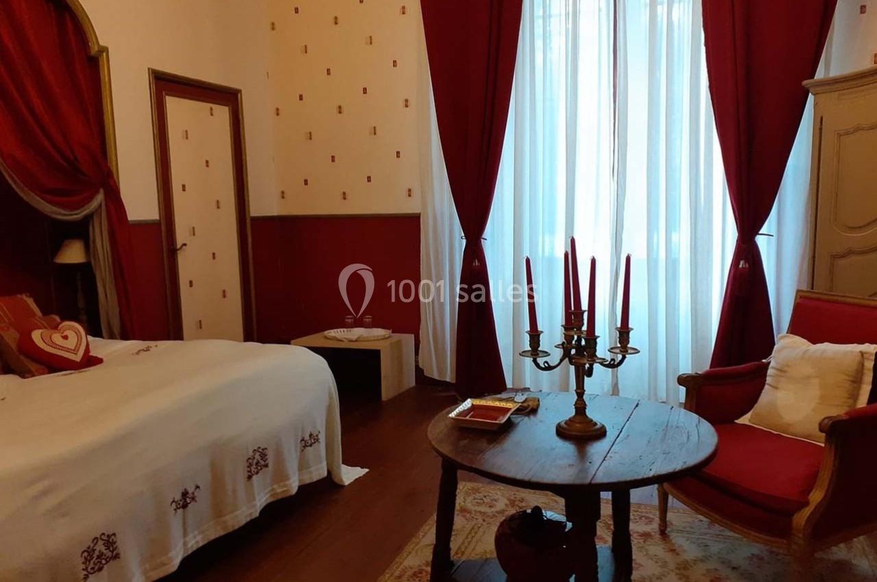 Chambre décorée de rouge avec lit, fauteuil, table ronde, chandelier et rideaux encadrant une grande fenêtre lumineuse.