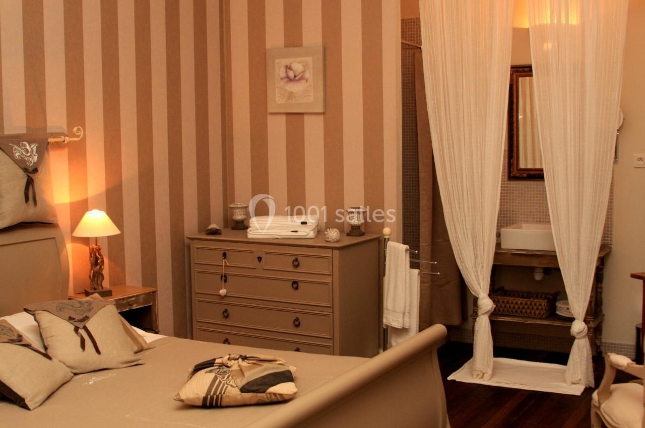 Chambre décorée dans des tons beiges avec lit, commode, lampes et un coin lavabo séparé par des rideaux blancs.