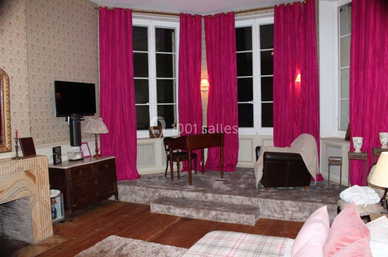 Salon avec parquet, cheminée, rideaux roses, meubles anciens, fauteuil et table en bois devant des fenêtres.