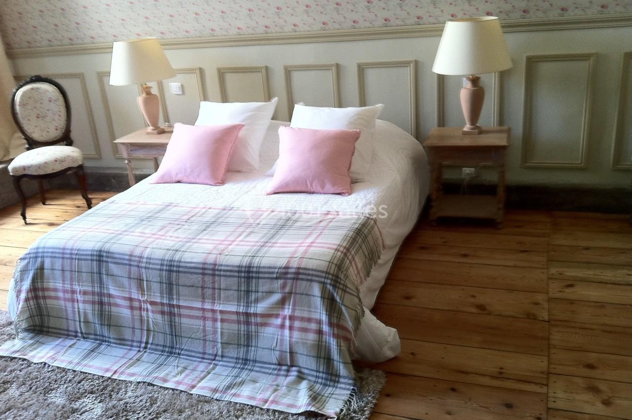 Lit double avec plaid à carreaux, coussins roses, tables de chevet avec lampes et chaise dans une chambre au sol en bois.