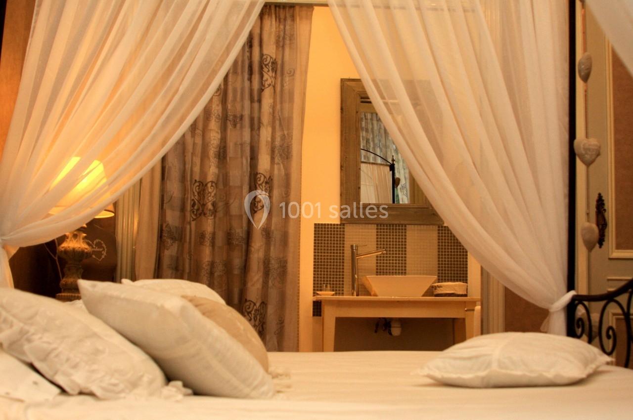 Lit à baldaquin avec voilages blancs, vue sur une coiffeuse et un lavabo dans une chambre chaleureusement éclairée.