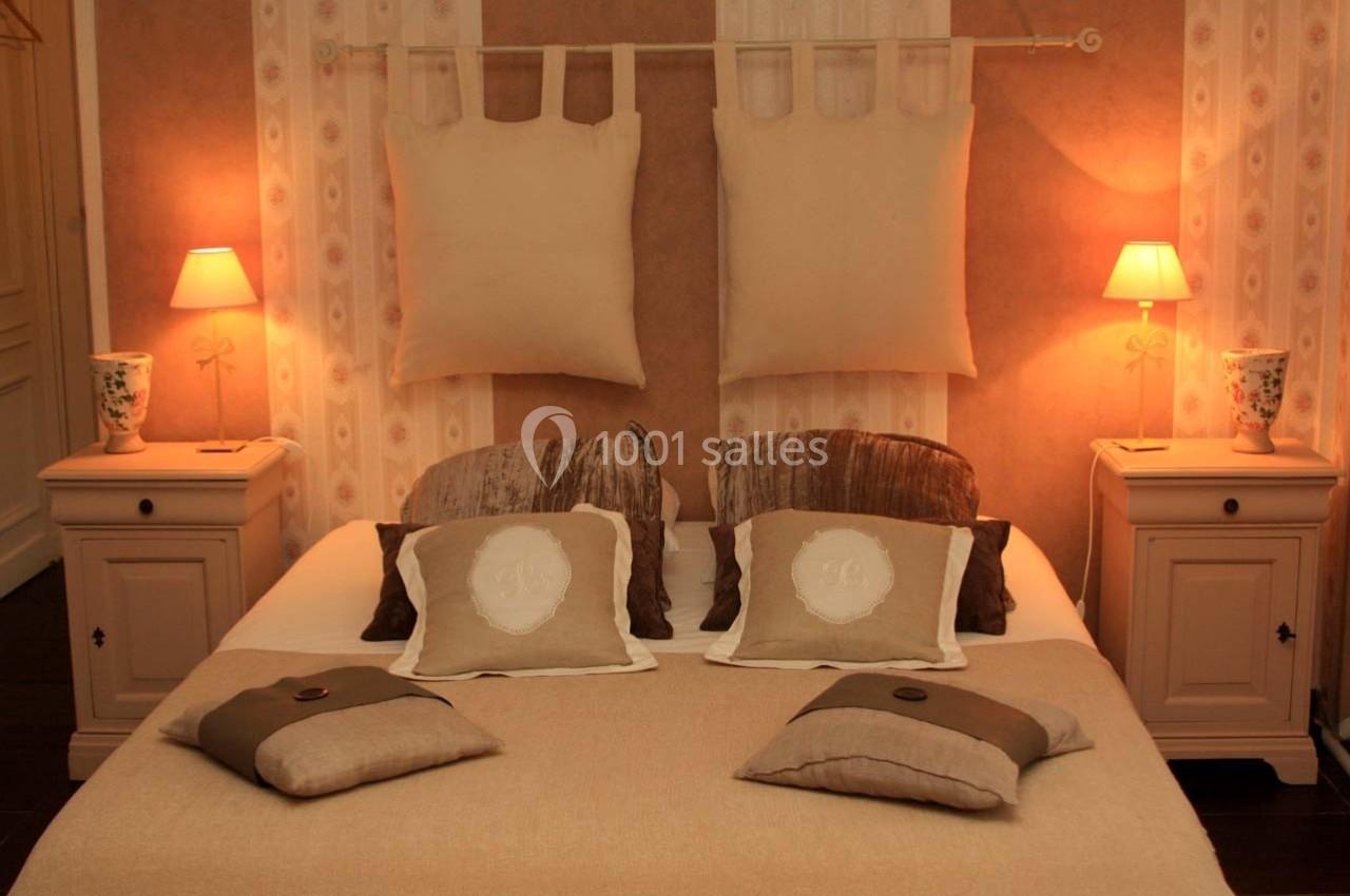 Lit double avec coussins beige et marron, encadré par deux lampes de chevet allumées sur des tables assorties.