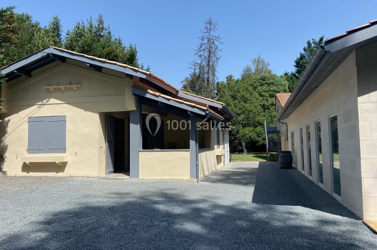 Deux bâtiments clairs avec toits en tuiles dans une cour gravillonnée, entourés d'arbres sous un ciel bleu.