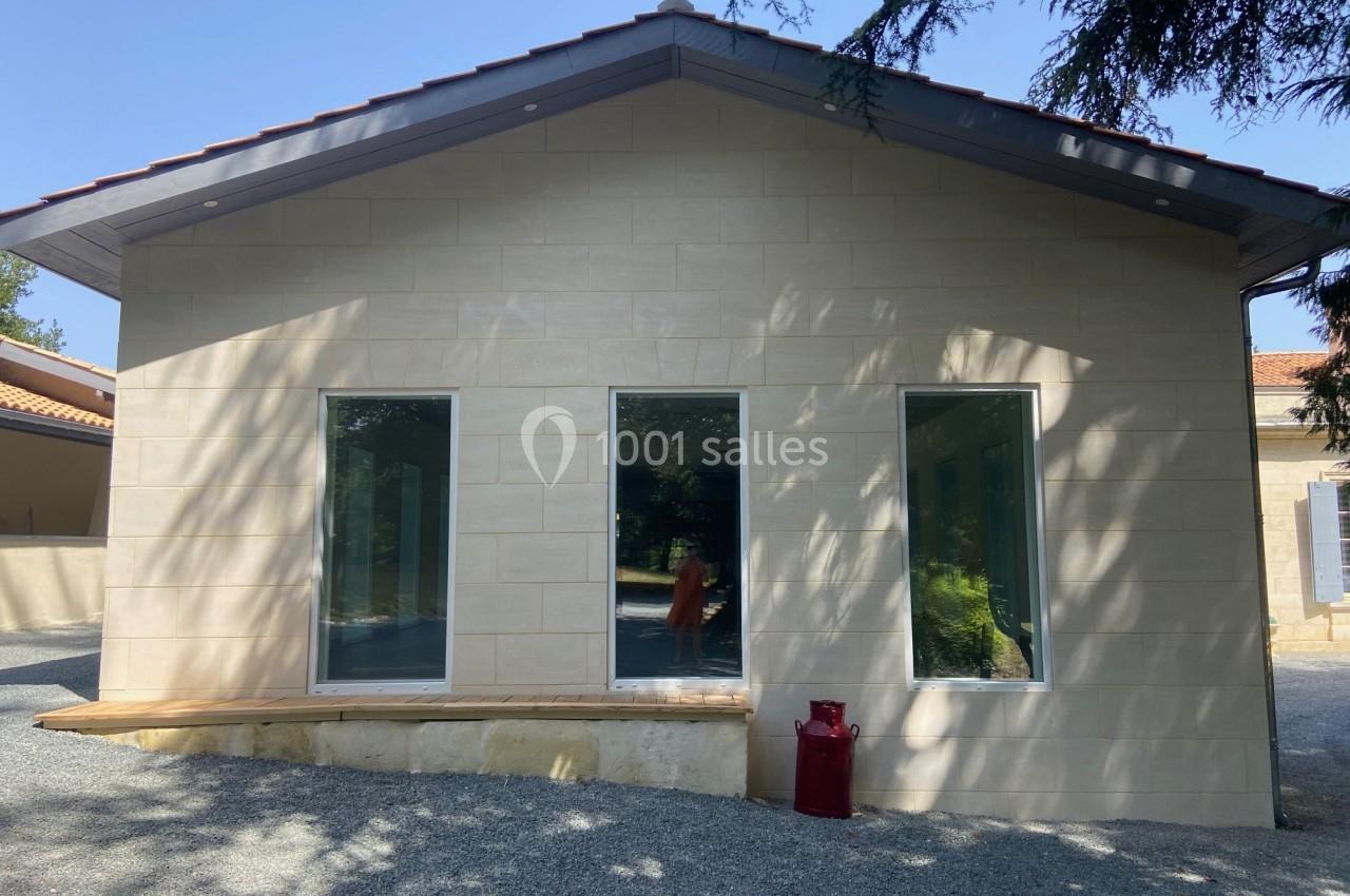 Façade d'un bâtiment en pierre claire avec trois grandes fenêtres, entourée de gravier et d'arbres.