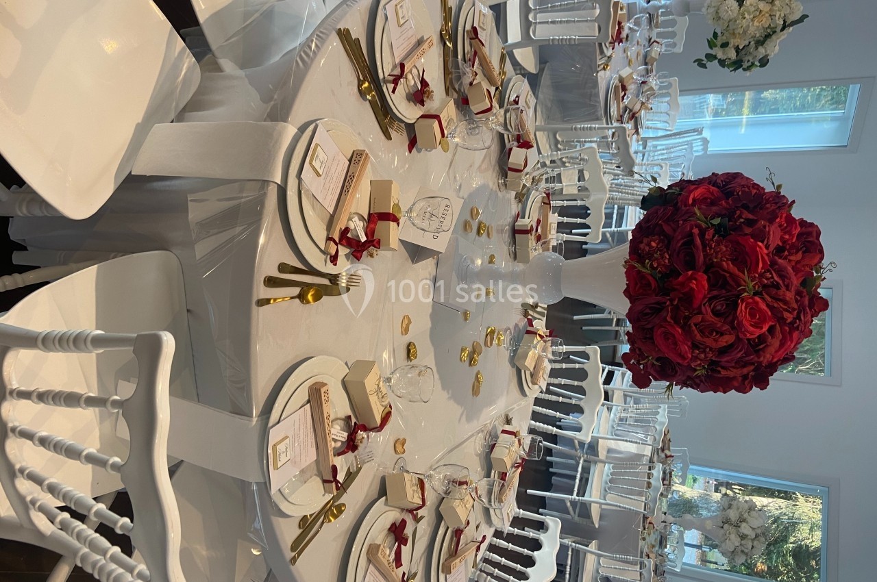 Table élégamment dressée avec nappes blanches, couverts dorés, menus et centre de table composé de roses rouges.