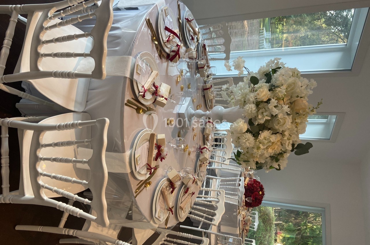 Table ronde élégamment dressée avec nappes blanches, couverts dorés, serviettes rouges et un centre floral blanc.