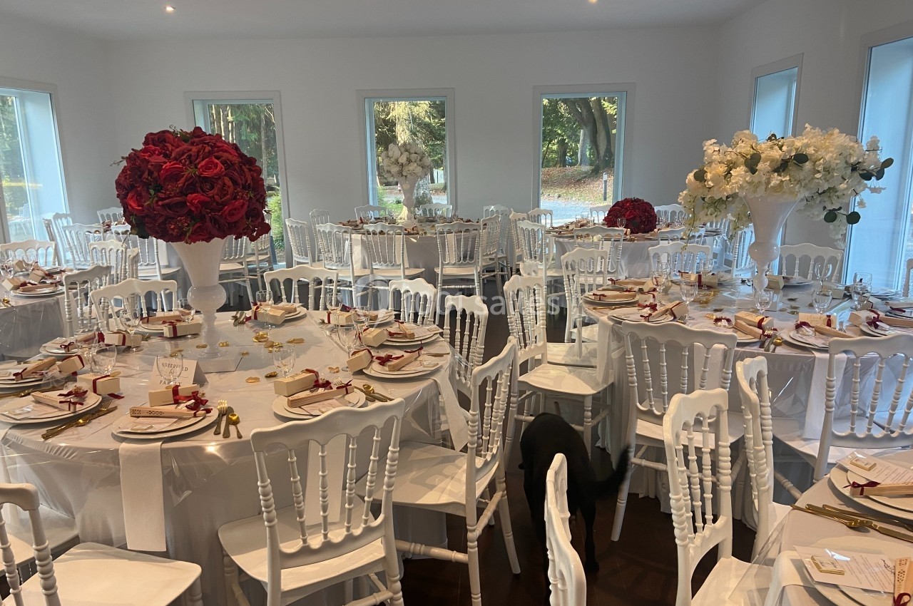 Salle de réception décorée avec des tables rondes, nappes blanches, chaises blanches et centres de table fleuris rouges et…
