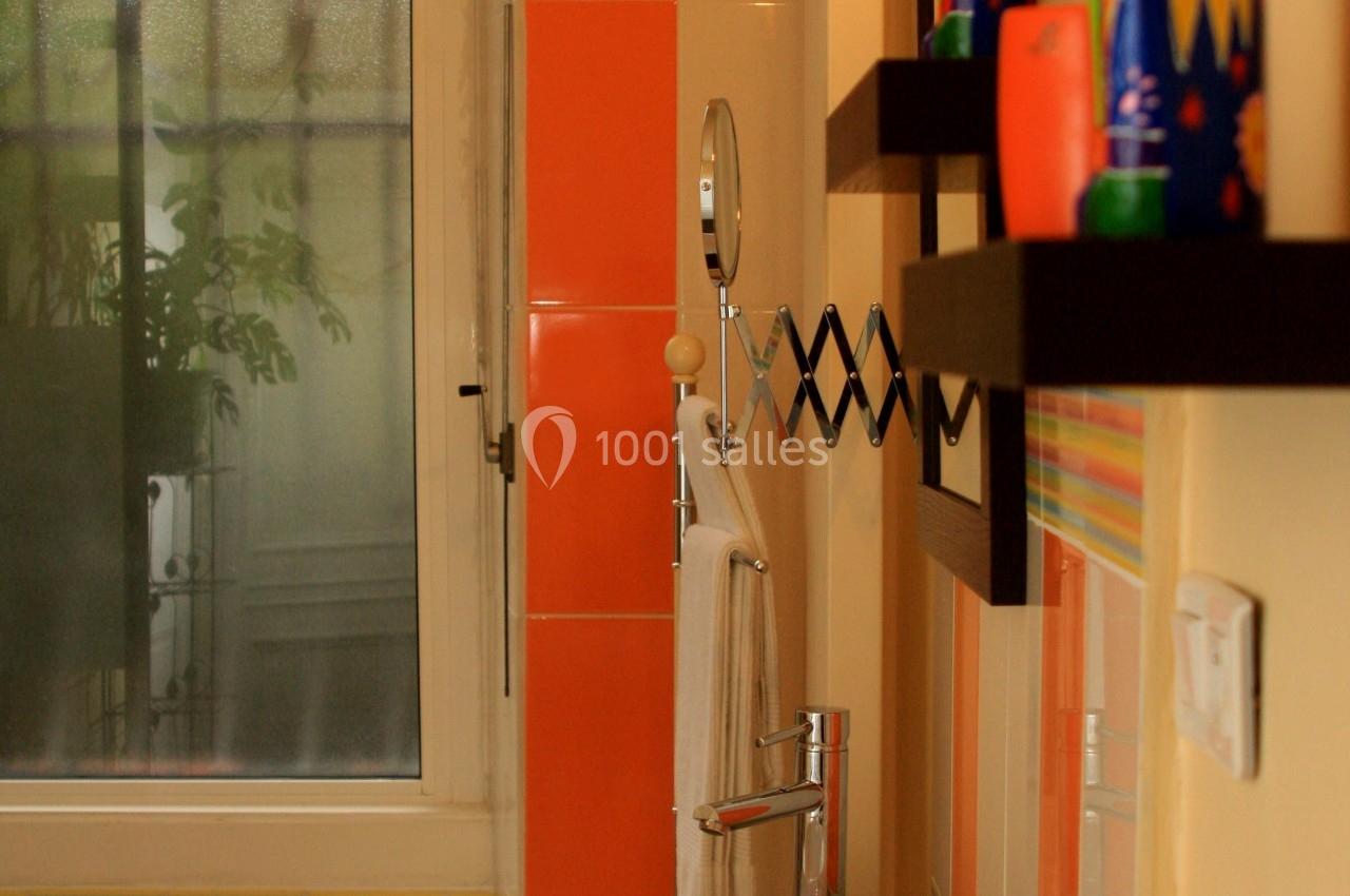Salle de bain colorée avec lavabo blanc, étagères décorées et fenêtre givrée laissant passer la lumière naturelle.