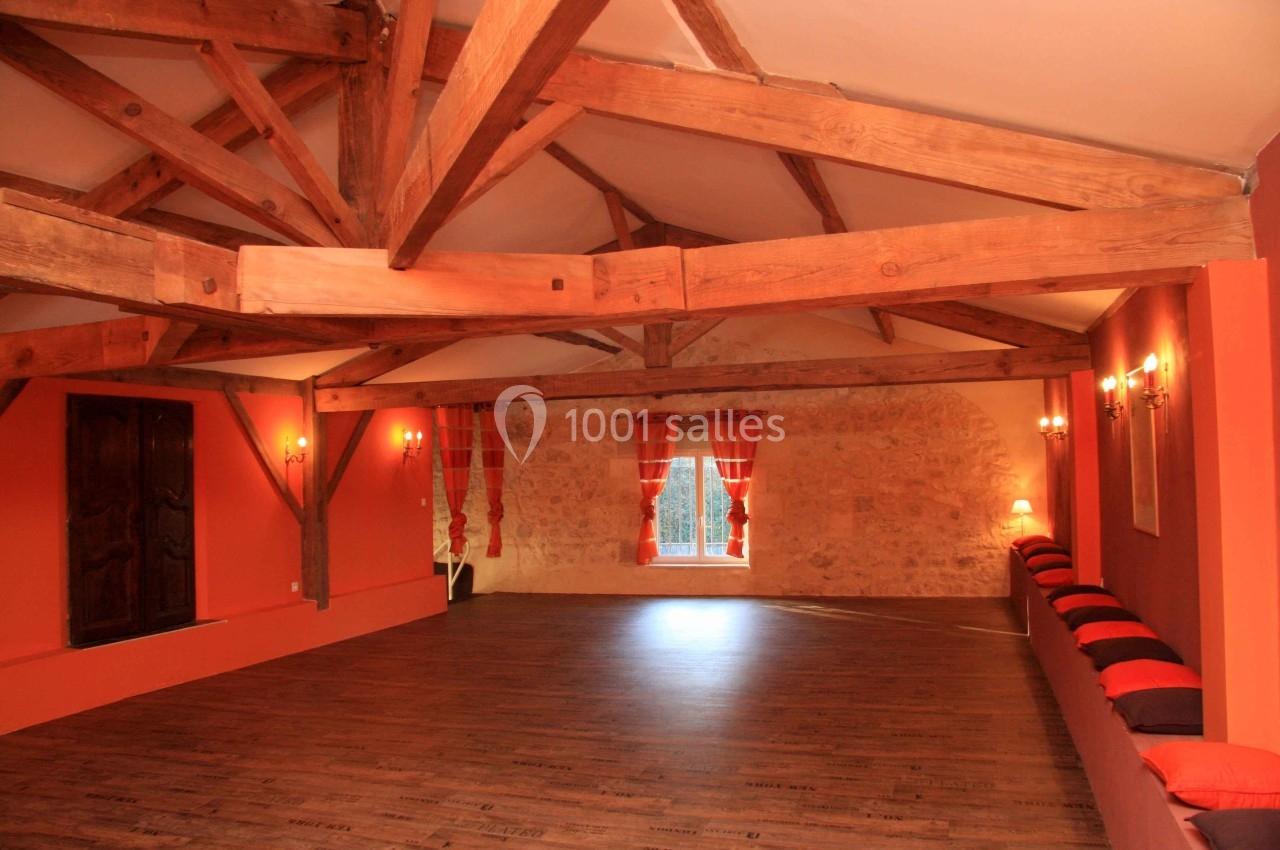Salle spacieuse avec poutres apparentes, parquet en bois, murs orange, rideaux rouges et banquettes avec coussins.