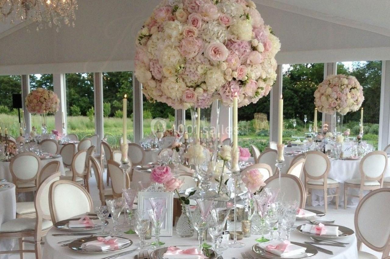 Salle de réception élégante avec tables dressées, décorations florales roses et blanches, et vue sur un jardin verdoyant.
