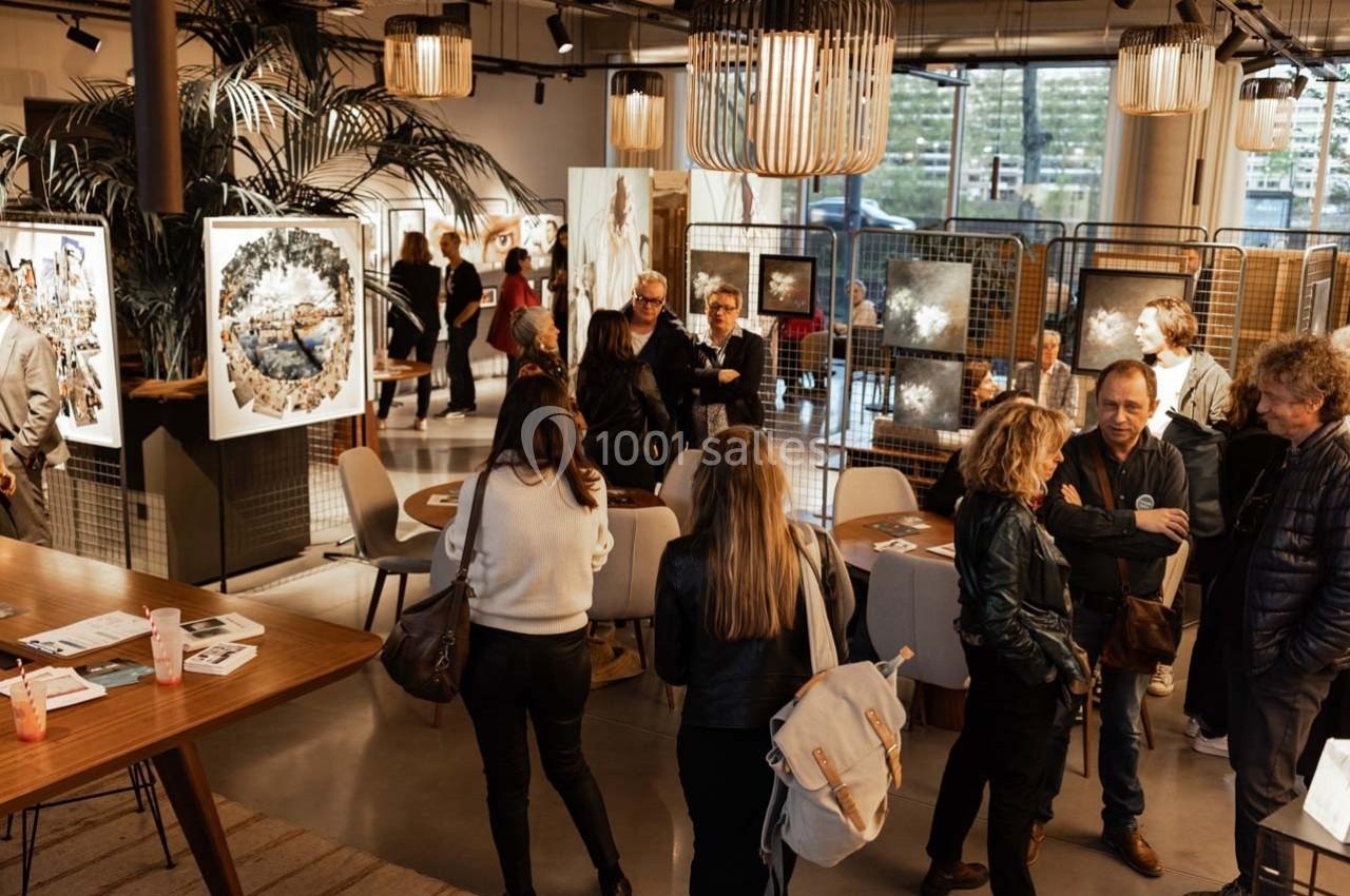 Des personnes visitent une exposition d'art dans un espace moderne avec des œuvres accrochées et des luminaires suspendus.