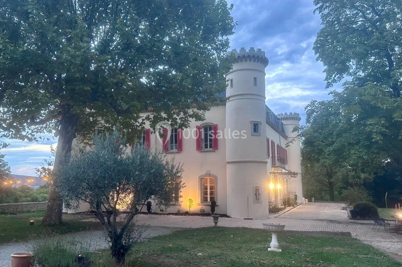 Château blanc avec tours rondes, volets rouges, entouré d'arbres et éclairé à la tombée de la nuit.