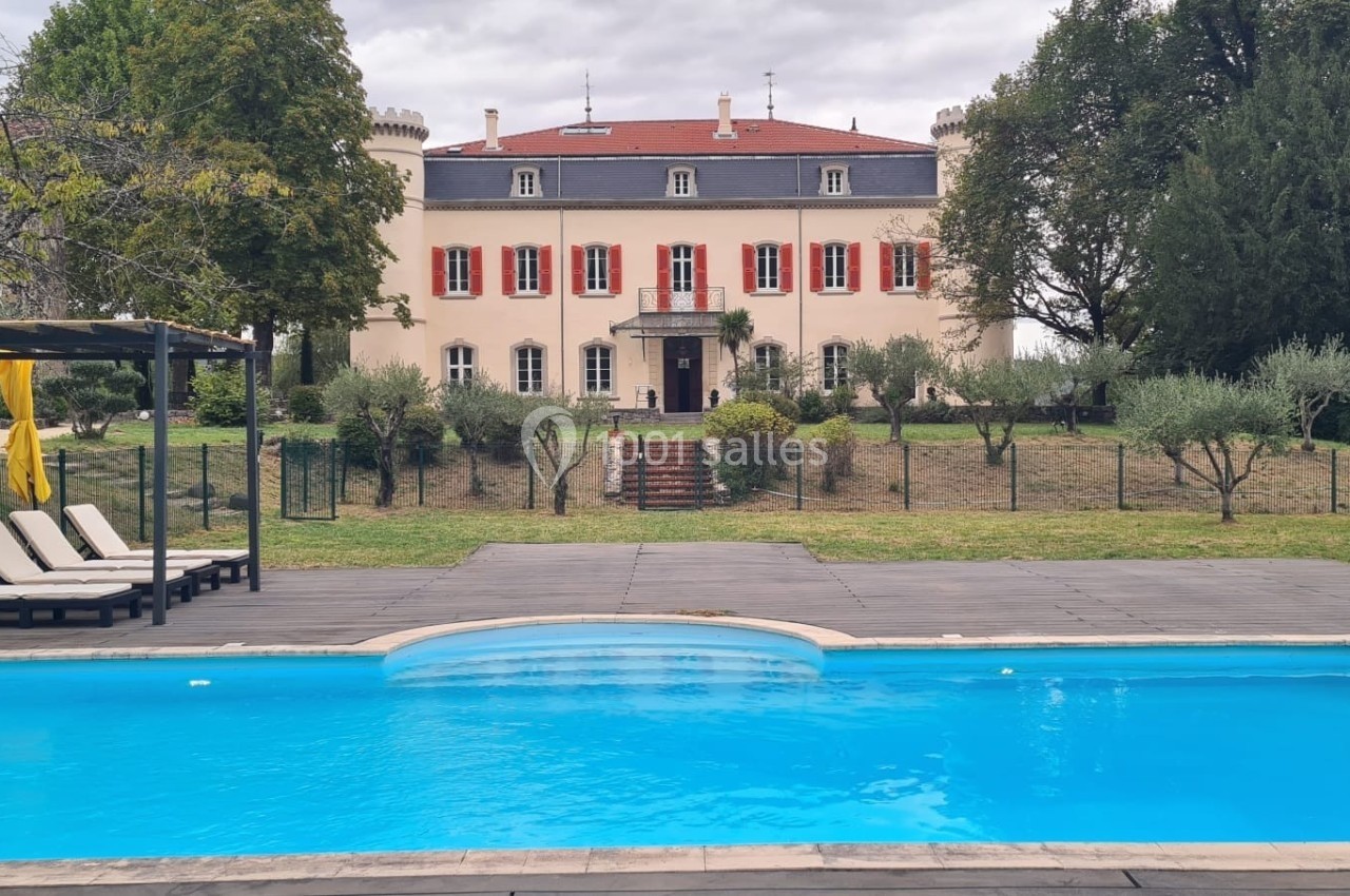 Piscine extérieure devant un grand bâtiment ancien avec volets rouges, entouré d'arbres et de chaises longues.