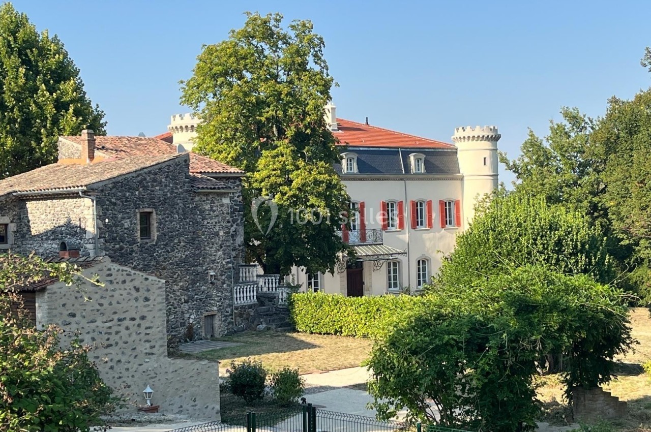 Bâtiment en pierre ancienne à gauche et château rénové avec tours et volets rouges à droite, entourés de verdure.