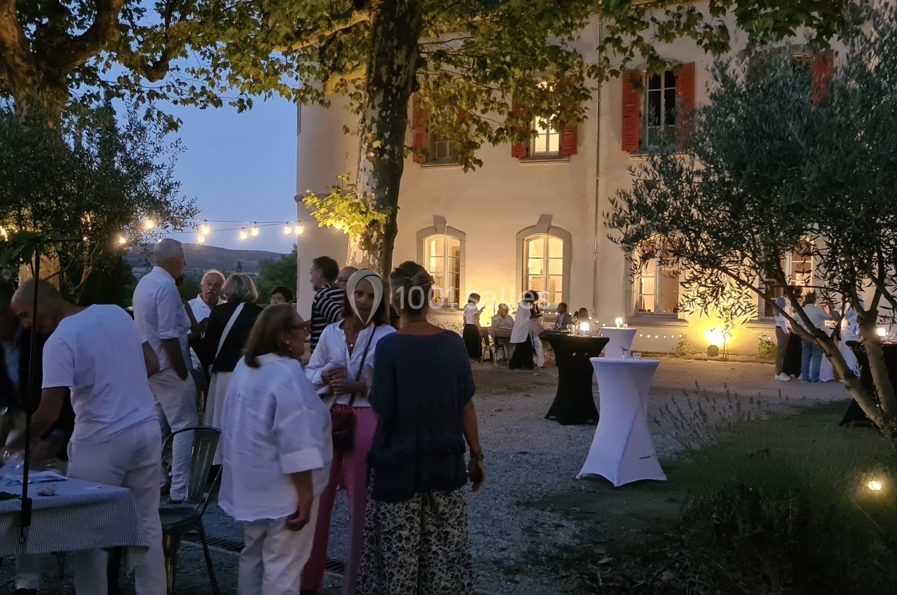 Groupe de personnes réunies en soirée dans un jardin éclairé, devant un bâtiment ancien avec des tables dressées.