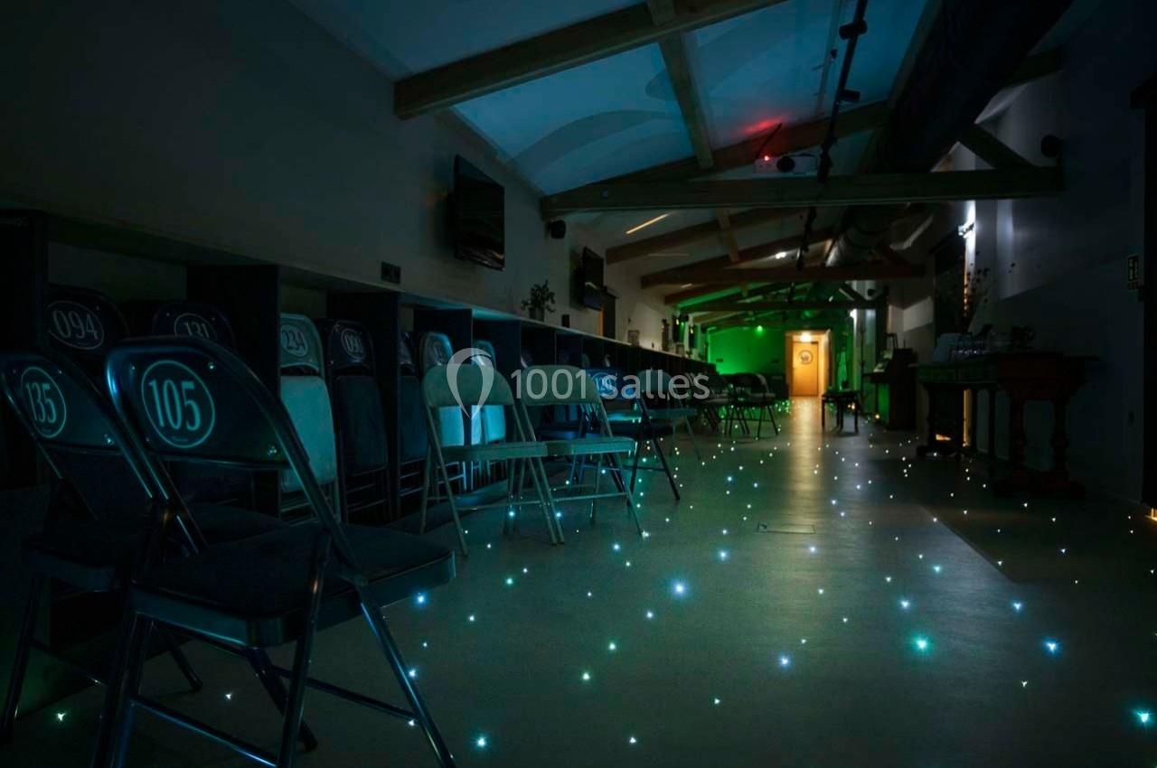 Salle sombre avec des chaises numérotées alignées, sol illuminé par des points lumineux et lumière verte au fond.