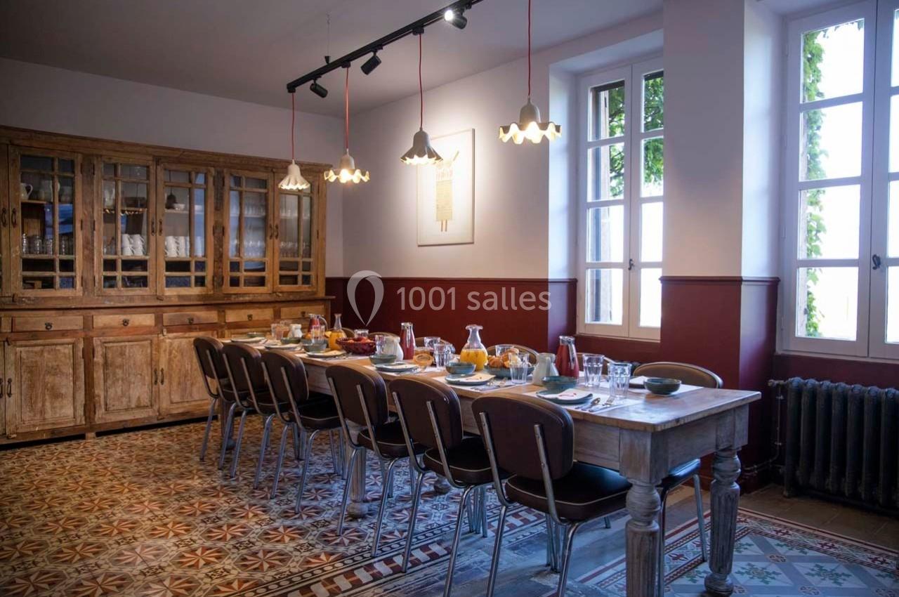 Salle à manger lumineuse avec une grande table en bois dressée pour un repas, entourée de chaises et éclairée par des…