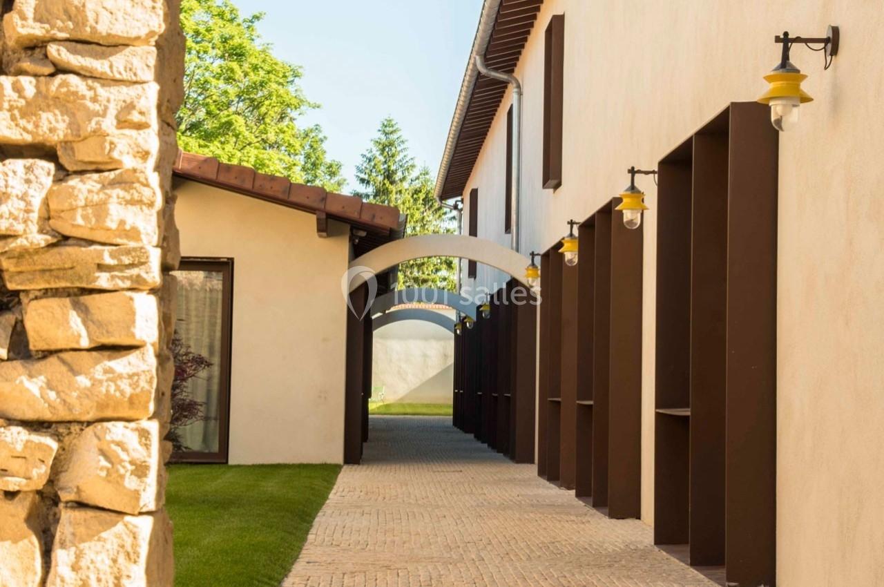 Passage extérieur pavé bordé de murs beige avec arches, éclairages muraux et verdure en arrière-plan.