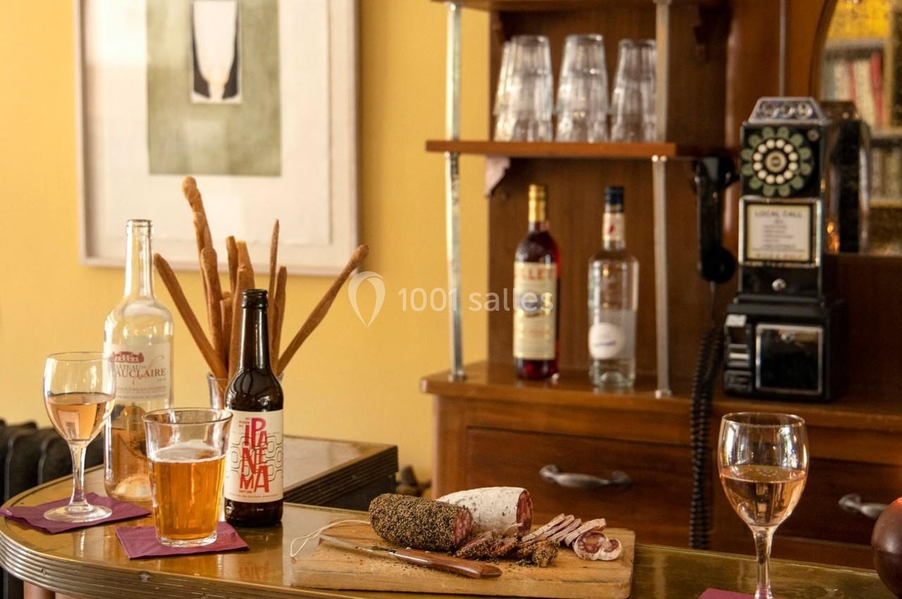 Planche de charcuterie avec verre de vin et bière sur un comptoir, entourée de bouteilles et d'un téléphone vintage.