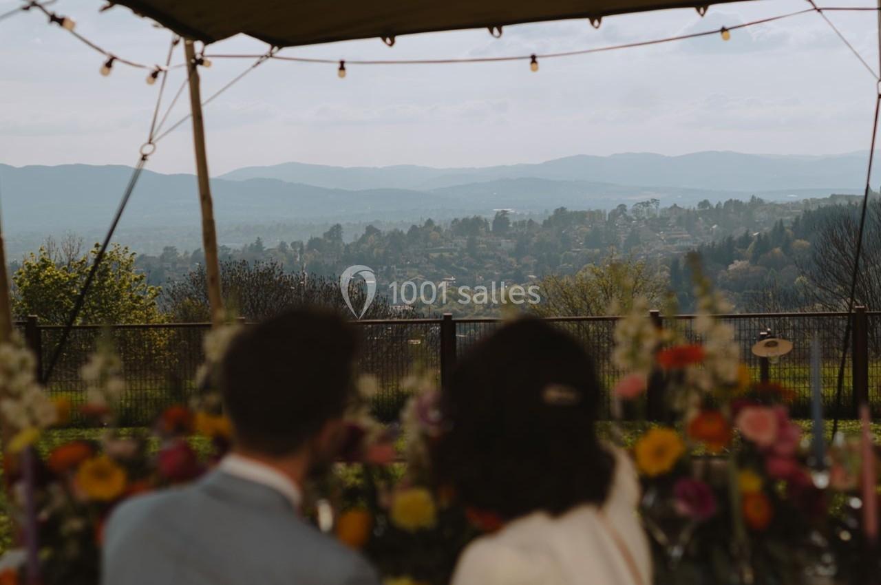 Un couple de dos observe un paysage vallonné sous une pergola décorée de fleurs colorées.